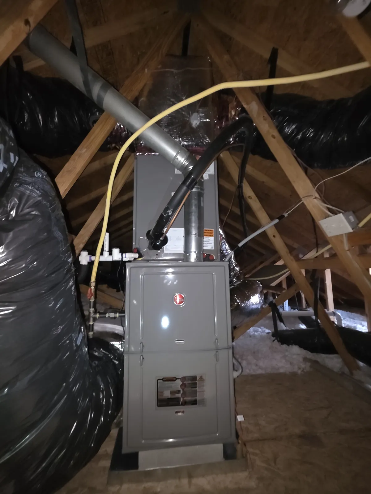 A new Rheem gas furnace standing upright in an attic with the coil cabinet above it, a metal flue pipe rising, a yellow gas line and PVC condensate fittings at its side, black-wrapped flex duct around it, on a North Texas job